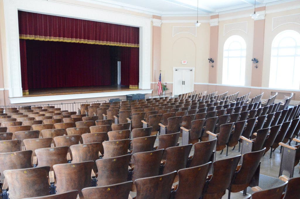 Auditorium view from the rear toward the stage, red curtain, arched windows, and restored wooden seating.