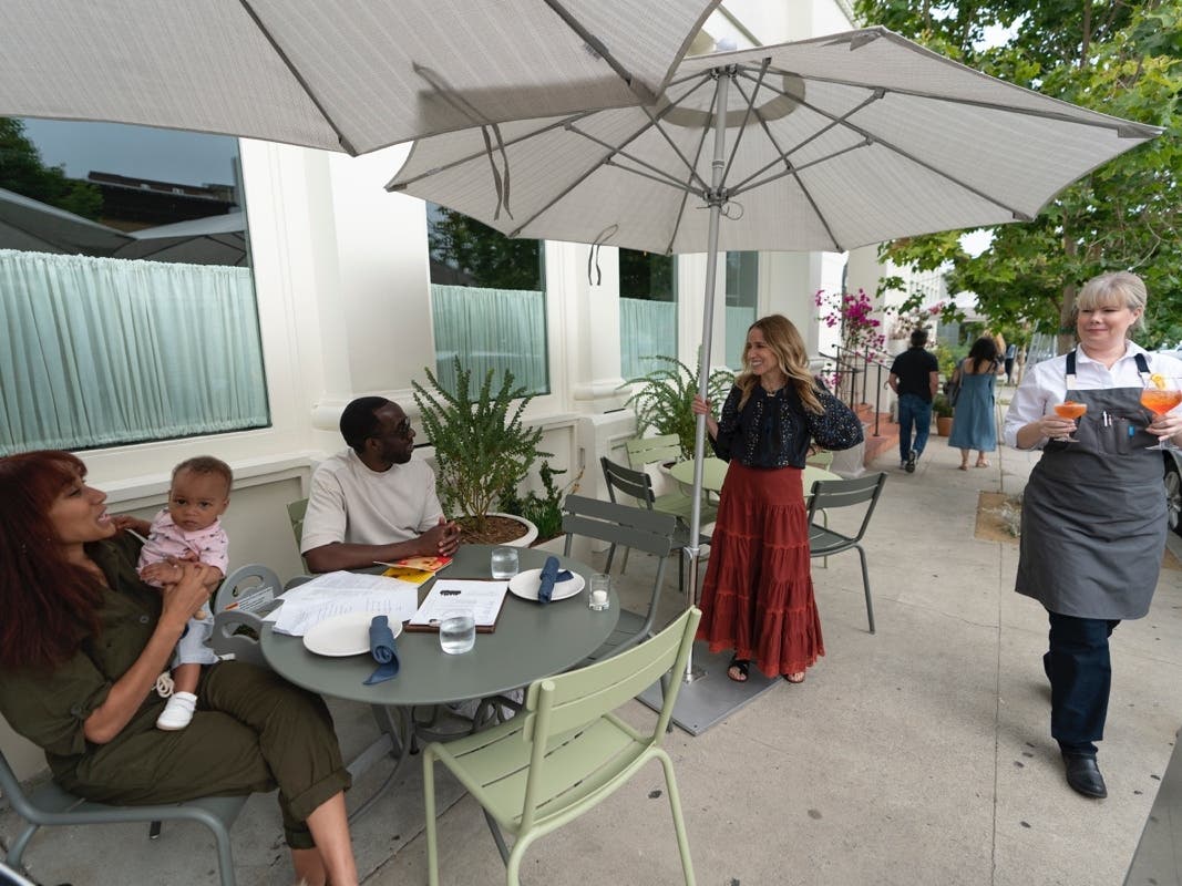 In this Saturday, June 19, 2021, photo, Caroline Styne, owner and wine director at The Lucques Group, standing under umbrella, welcomes back regular customers, Chris Anokute with his wife Jasmine and their 9-month-old son, Phoenix, at the A.O.C. 