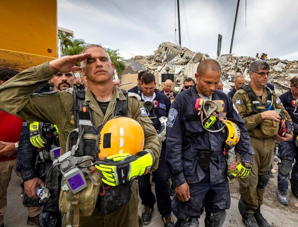 A member of the Israeli search and rescue team, left, salutes in front of the rubble that once was Champlain Towers South during a prayer ceremony held Wednesday.