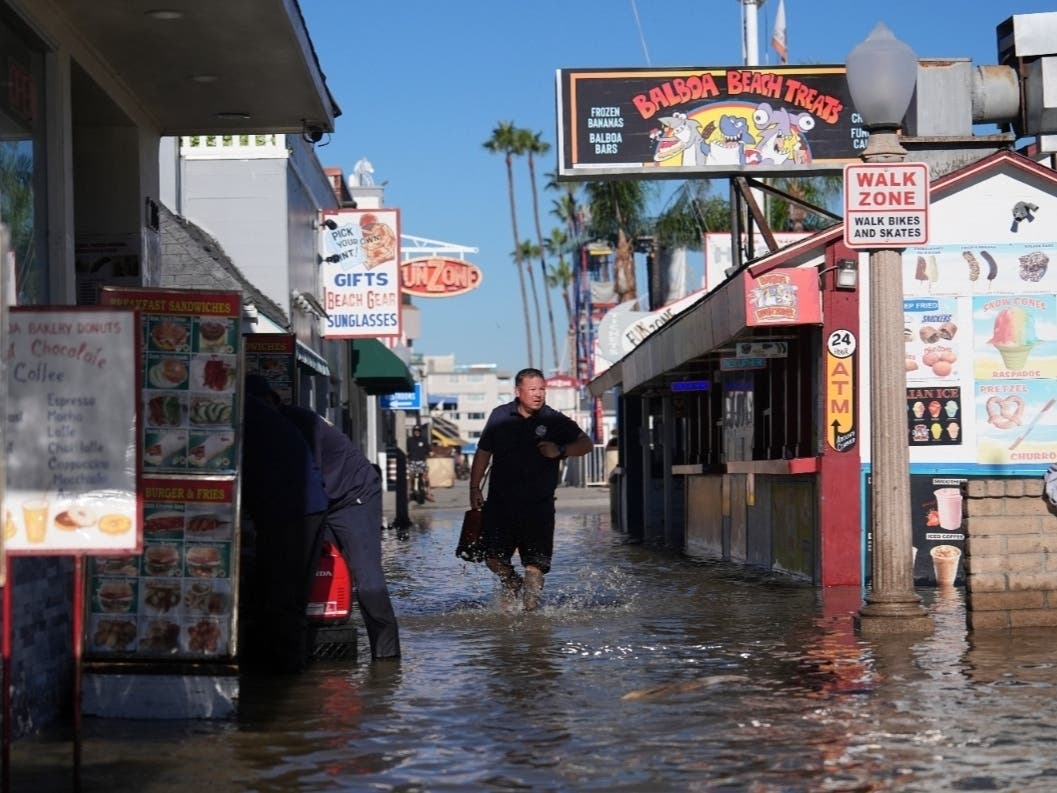 A worker wades through floodwaters along a row of beachfront businesses in Newport Beach on Monday, Oct. 26, 2026, as record-breaking high tides inundated coastal communities across California.