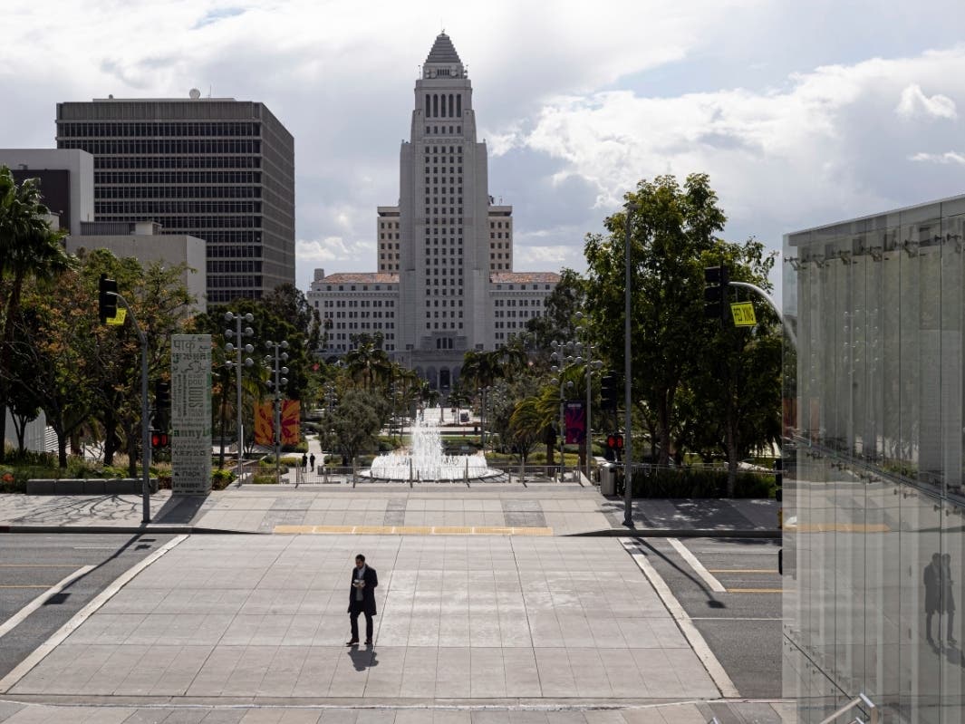 A lone pedestrian crosses Grand Avenue with Los Angeles city hall as a backdrop in downtown Los Angeles on Thursday, March 19, 2020.