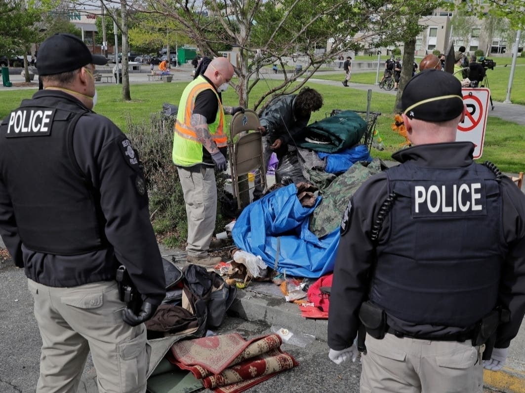 Seattle Police officers and other workers look on as a man packs up his belongings from a homeless encampment being cleared Monday, May 4, 2020, at Ballard Commons Park in Seattle.