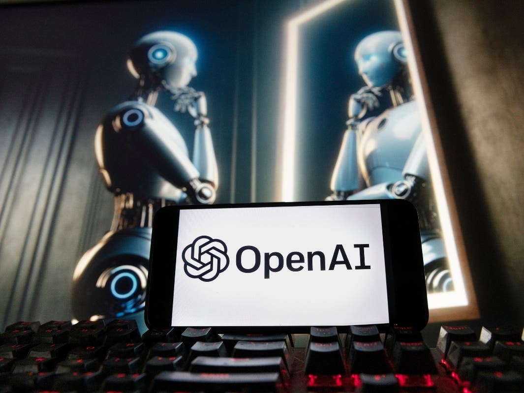 The OpenAI logo is displayed on a cellphone with an image on a computer monitor generated by ChatGPT's Dall-E text-to-image model.