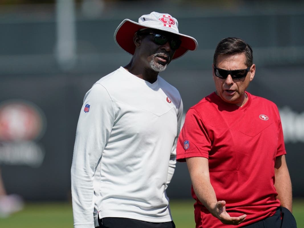 San Francisco 49ers assistant coach Johnny Holland, left, talks with broadcaster Greg Papa as players takes part in drills at the NFL football team's practice facility in Santa Clara, Calif., Saturday, July 30, 2022