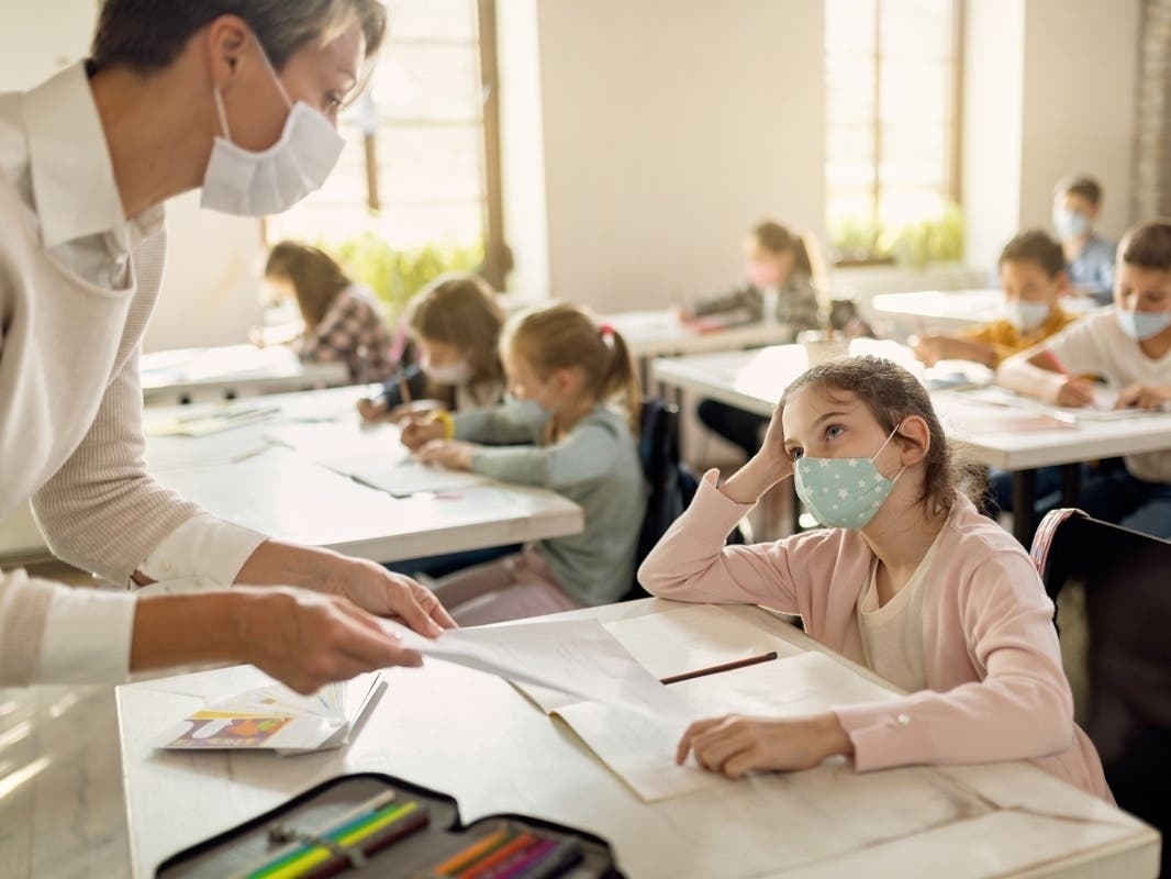 Schoolgirl with face mask talking to a teacher while receiving test results in the classroom.