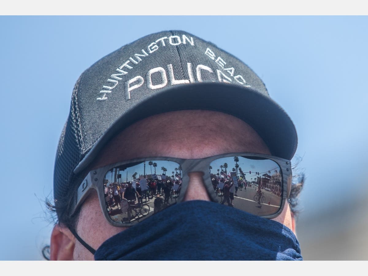A police officer wears a face scarf as a preventive measure against the spread of the coronavirus while he watches protestors gathering in a demonstration on May 1, 2020 in Huntington Beach, California.