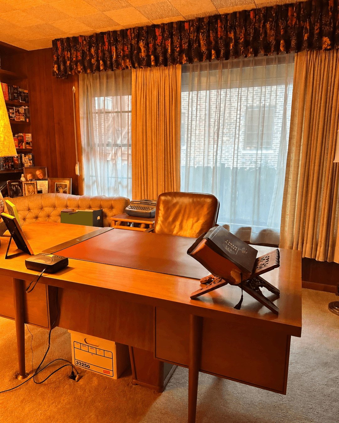 Wood-paneled office with a large wooden desk, leather chair, bookshelves, family photographs, and gold curtains.