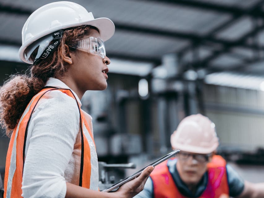 Female industrial engineer wearing a white helmet while standing in a heavy industrial factory behind she talking with workers, Various meta