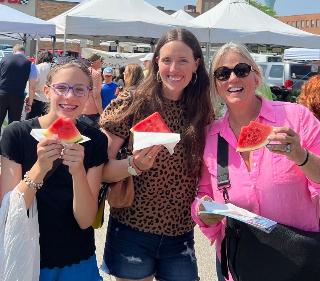 Free slices of watermelon will be available at Northbrook Farmers Market Opening Day festivities, Wednesday, June 19.  Enjoying watermelon at last year’s opening (left to right) Maddie and Marie Junkmann, Northbrook, and Tamara Taylor Holmes, Glenview.