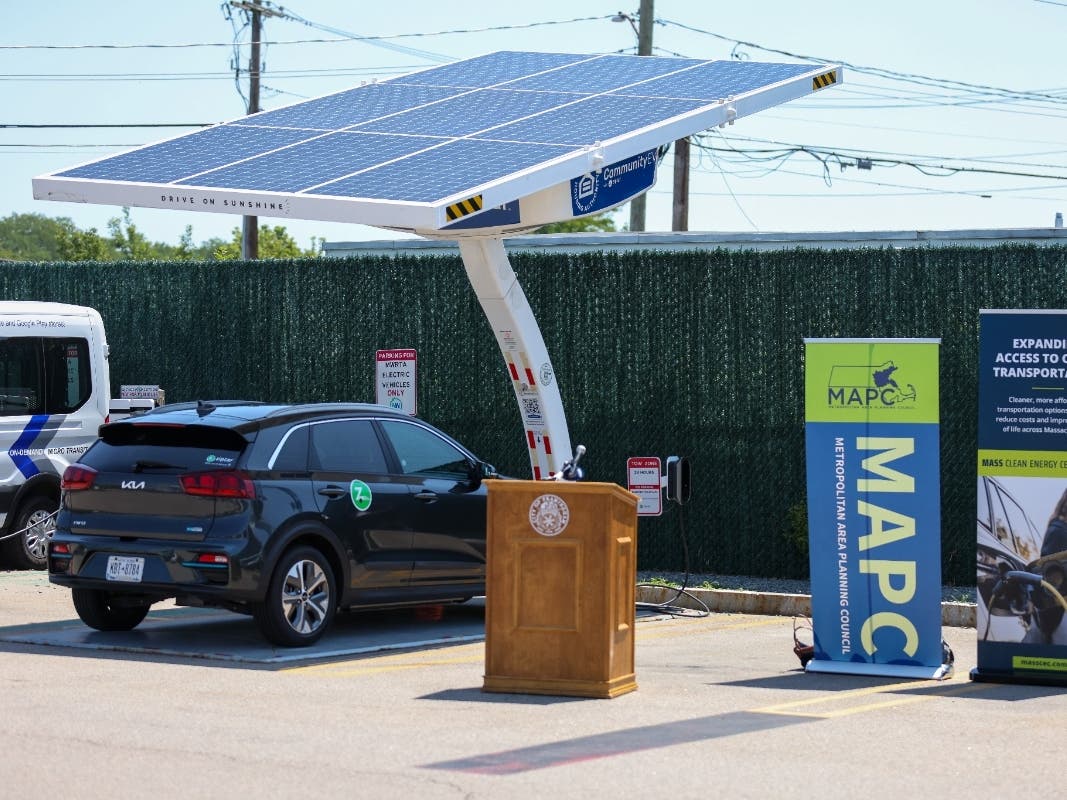 A Zipcar electric vehicle sits under a standalone, off-grid solar charger in the MetroWest RTA parking lot. A podium and banners from MAPC and MassCEC are set up in front of the vehicle, for a press conference launching the CommunityEV Carshare program.