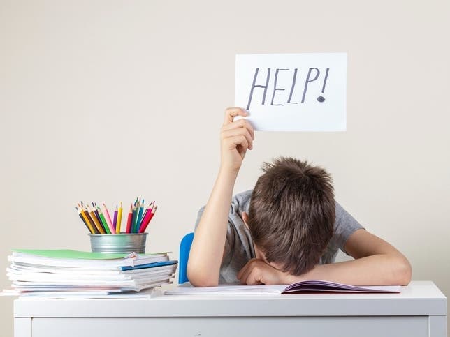 Sad tired frustrated boy sitting at the table with many books and holding paper with word Help. Learning difficulties, education concept.