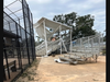 A view from the left field outside the ball diamond fence toward the small bleachers and grandstand shows pieces of sheet metal. A well-watered infield Inside the fence looks as if it only needs chalk lines applied to be ready for the next game.