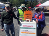 The three market masters of the Reston Farmers Market, from left, Keith Strange, and John and Fran Lovaas, pose for a photo Saturday next to a sign at the entrance of the market telling people to wear masks. After the photo, they put their masks back on.