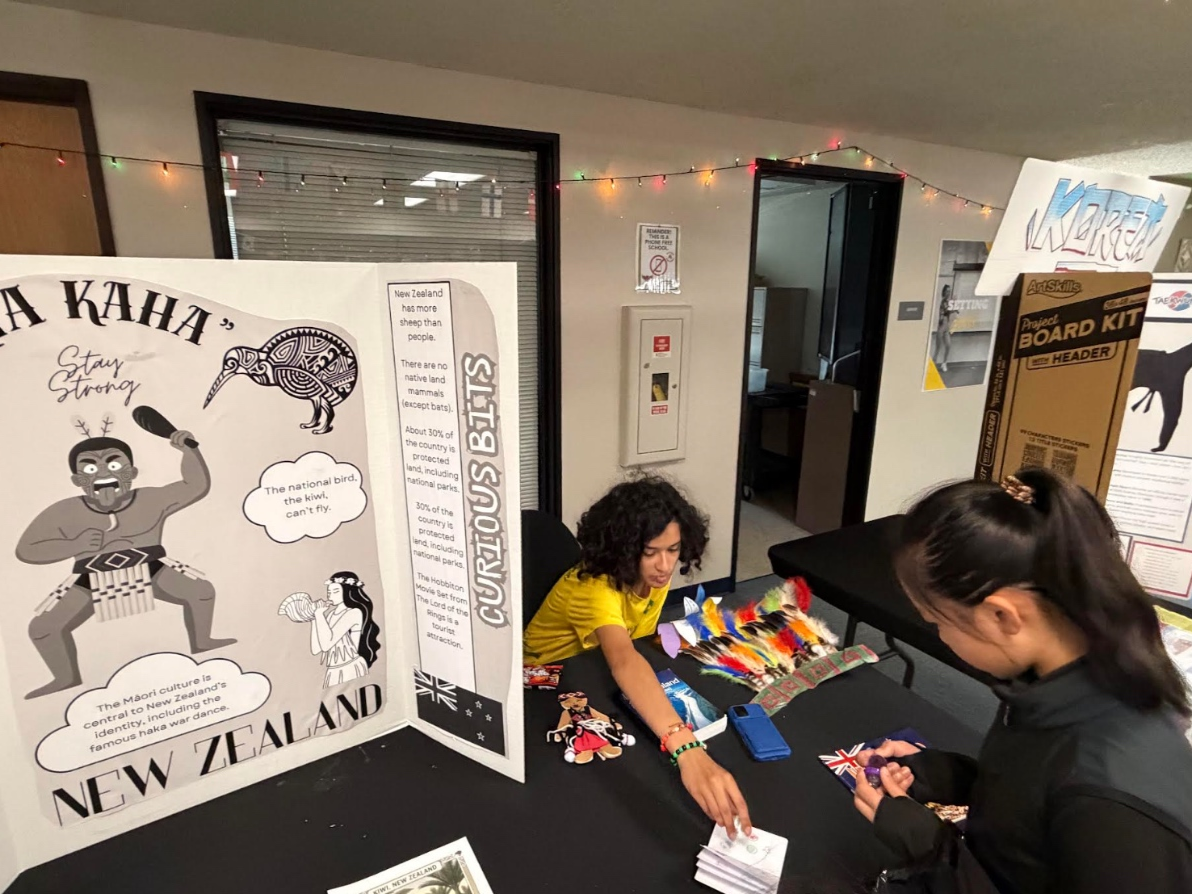 At the New Zealand table, a student leadership member helps another student add a country stamp to their passport.