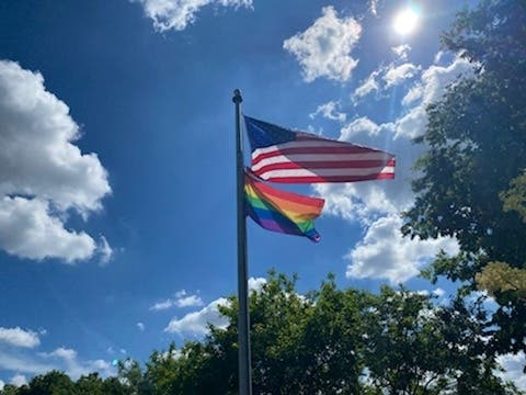 A Pride flag is shown flying with the U.S. Flag at the Rotary Village Green.