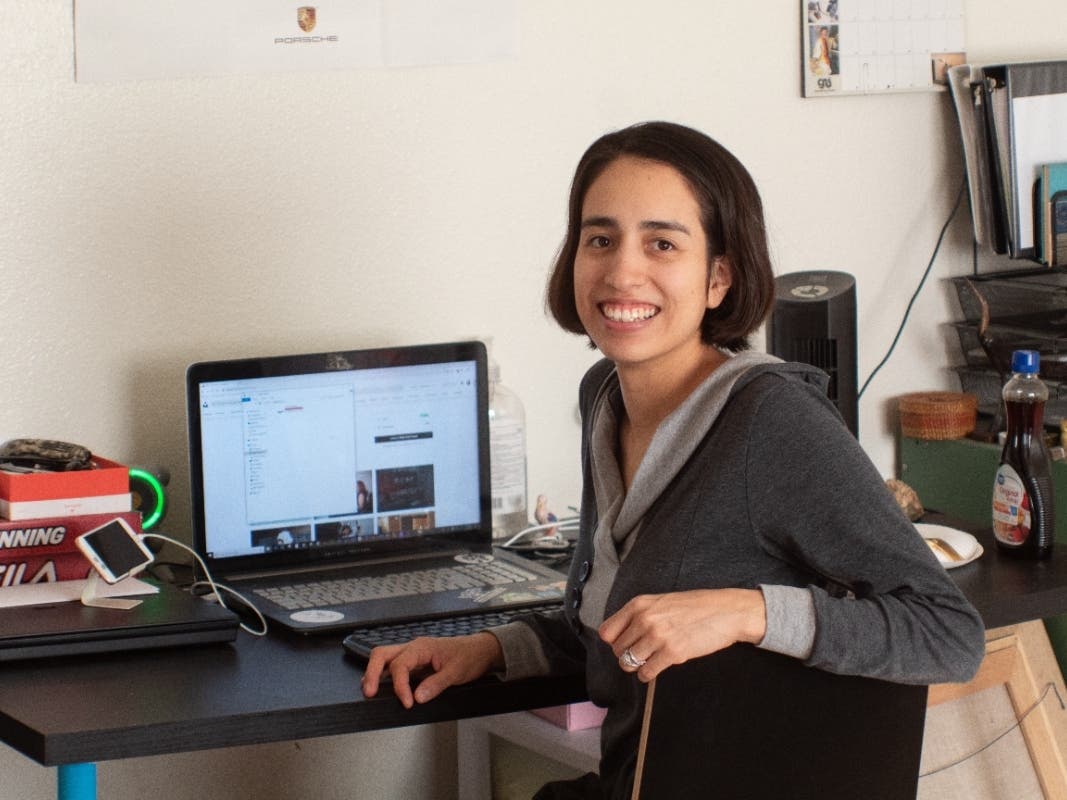A woman with brown hair sits at her desk with her laptop open, smiling at the camera