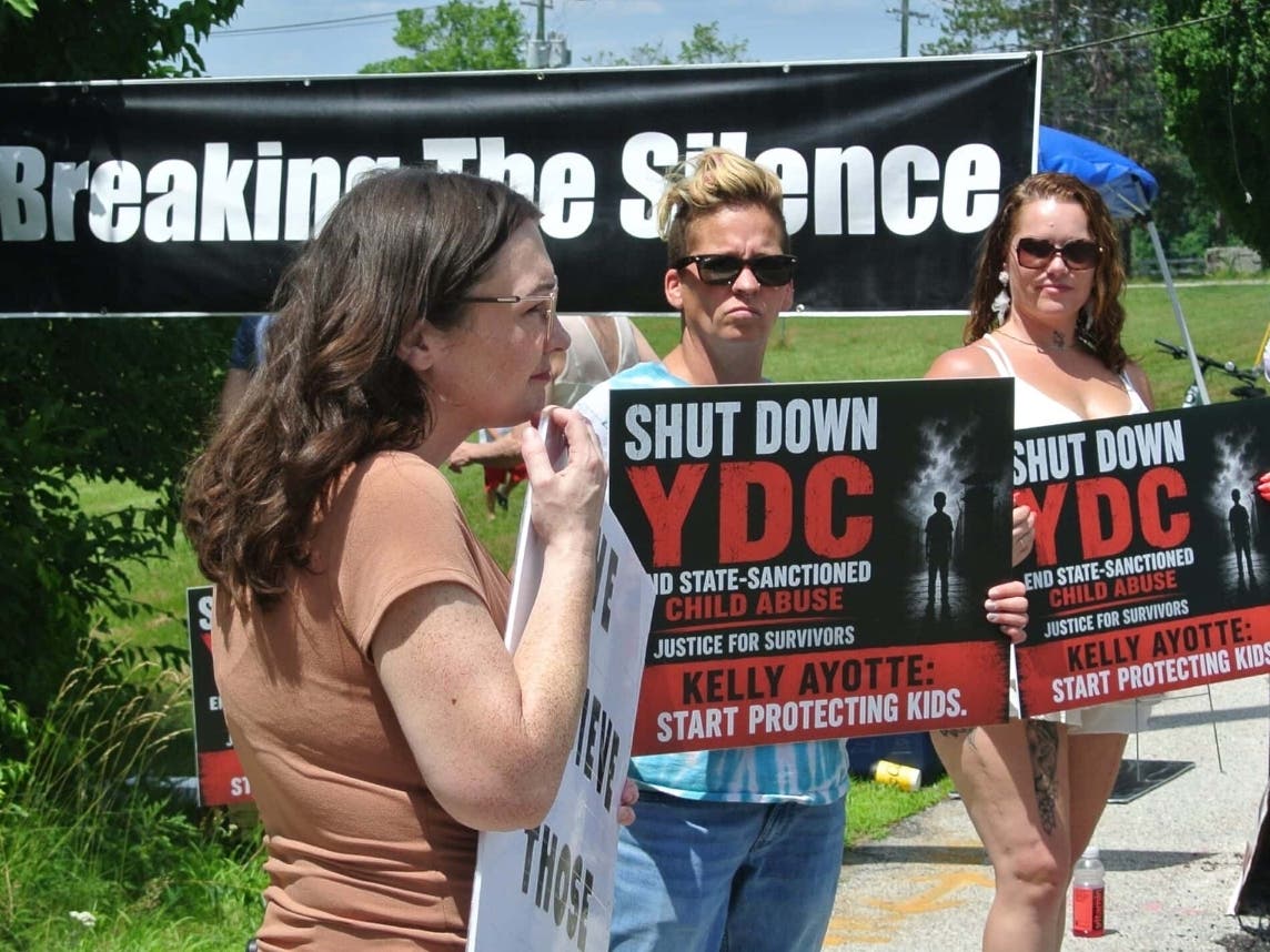 Survivor Michaela Jancsy, protestor Lela Bee and survivor Natasha Maunsell hold signs during their protest outside the Sununu Youth Services Center in Manchester.