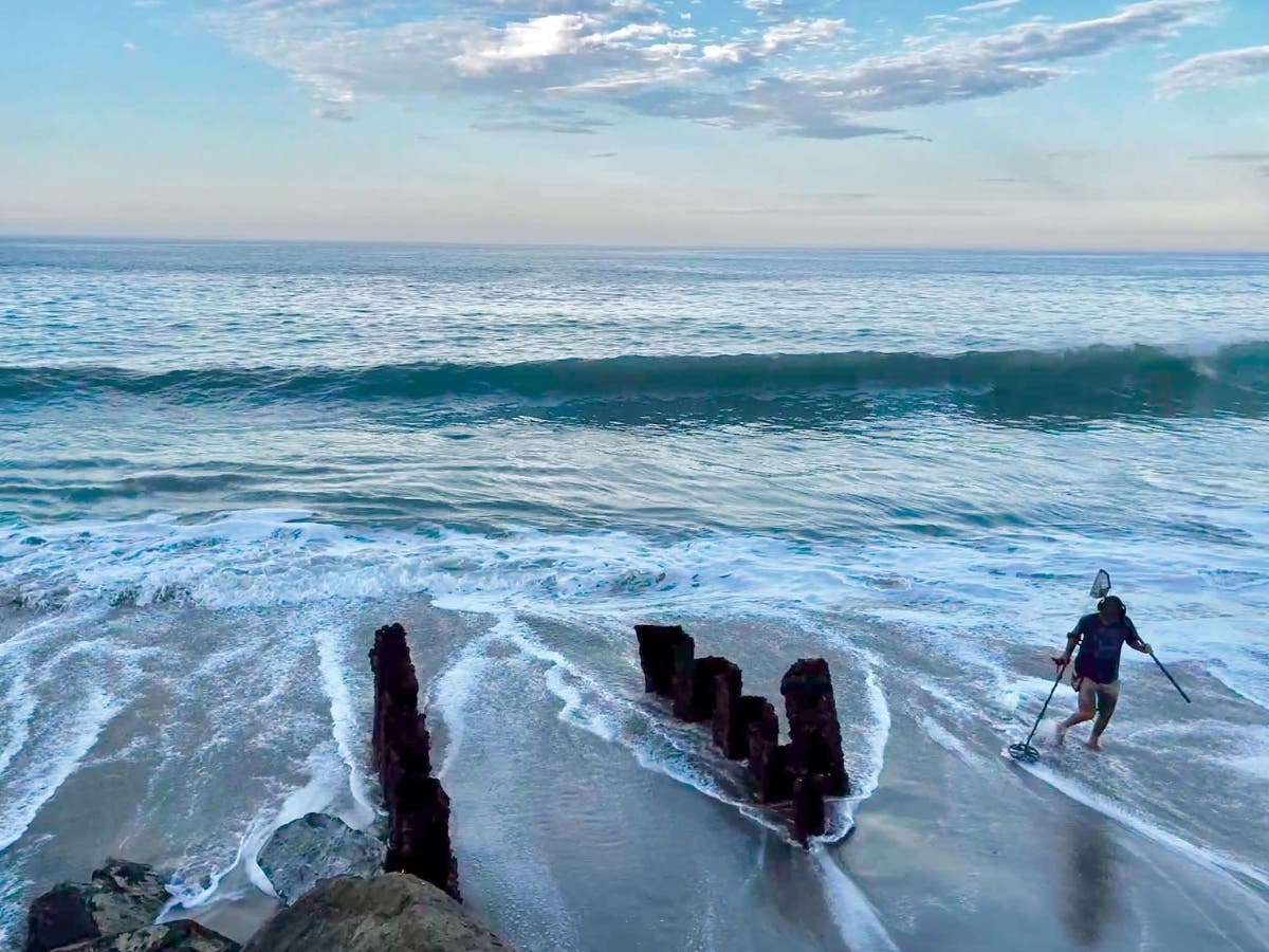 A man with a metal detector walks the shoreline at Aliso Beach, where waves have eroded away much of the sand, exposing old construction.
