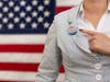 A woman standing in front of an American flag points to a "Vote" button on her lapel.