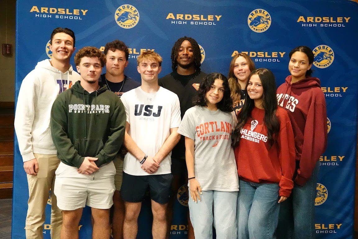 A group of student-athletes wearing collegiate gear smiling in front of a blue Ardsley backgdrop.