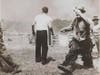 A man dressed in a clown costume carries a bucket in his right hand and shields his eyes with his left hand. This is Emmett Kelly, Sr., perhaps America's most famous circus clown.