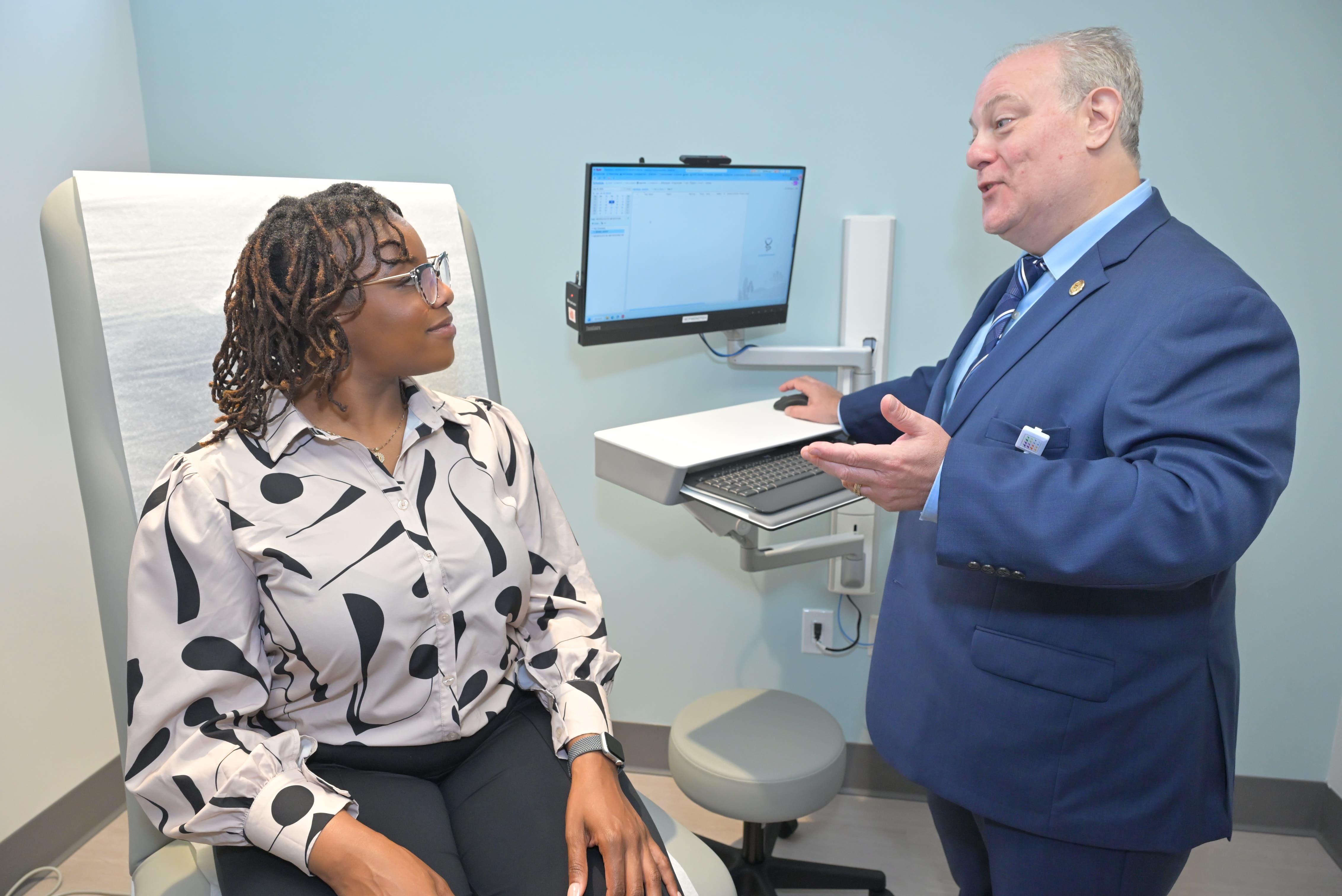 A happy male surgeon wearing a dark blue suit stands at a computer while speaking with a young female patient sitting on an exam room chair.