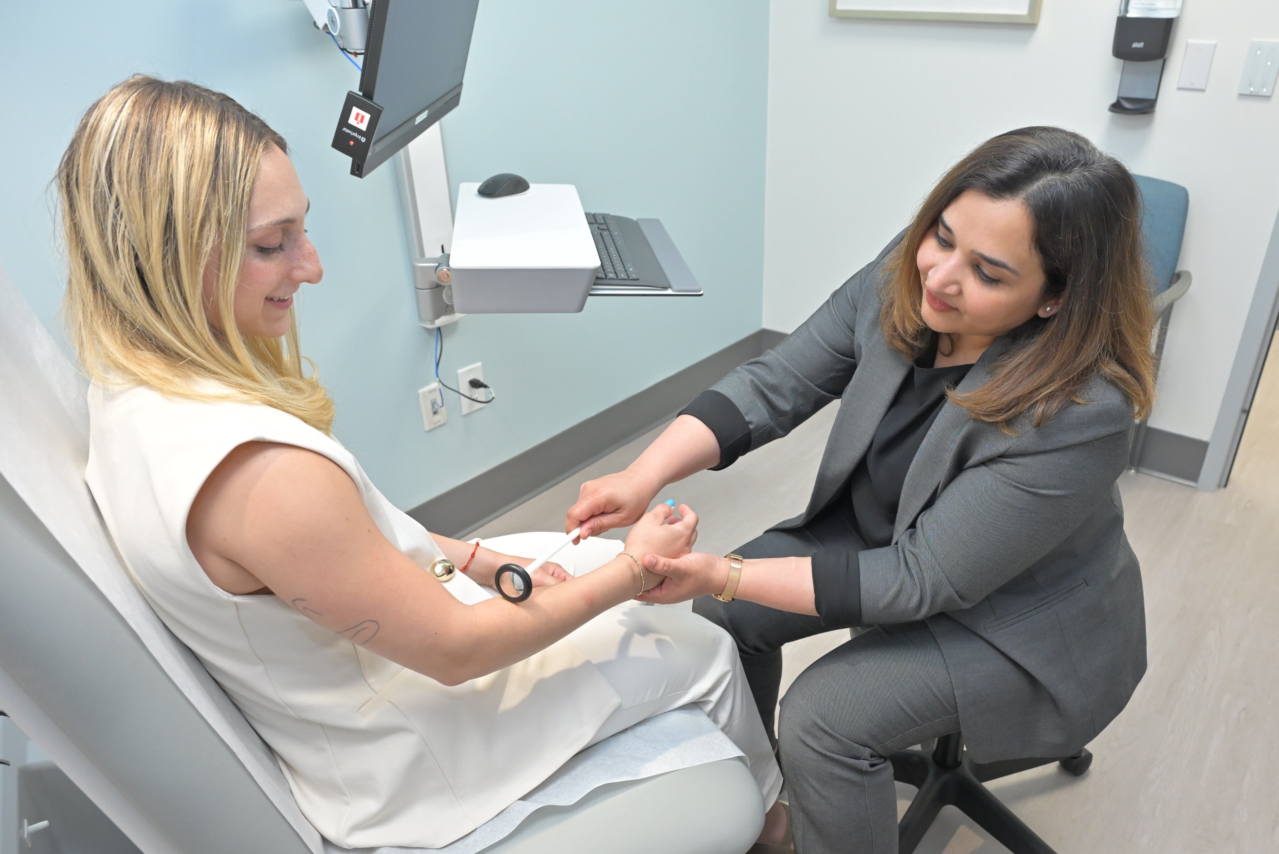 A female neurologist wearing a gray suit performs tests on a young blonde female patient sitting in an exam room chair.