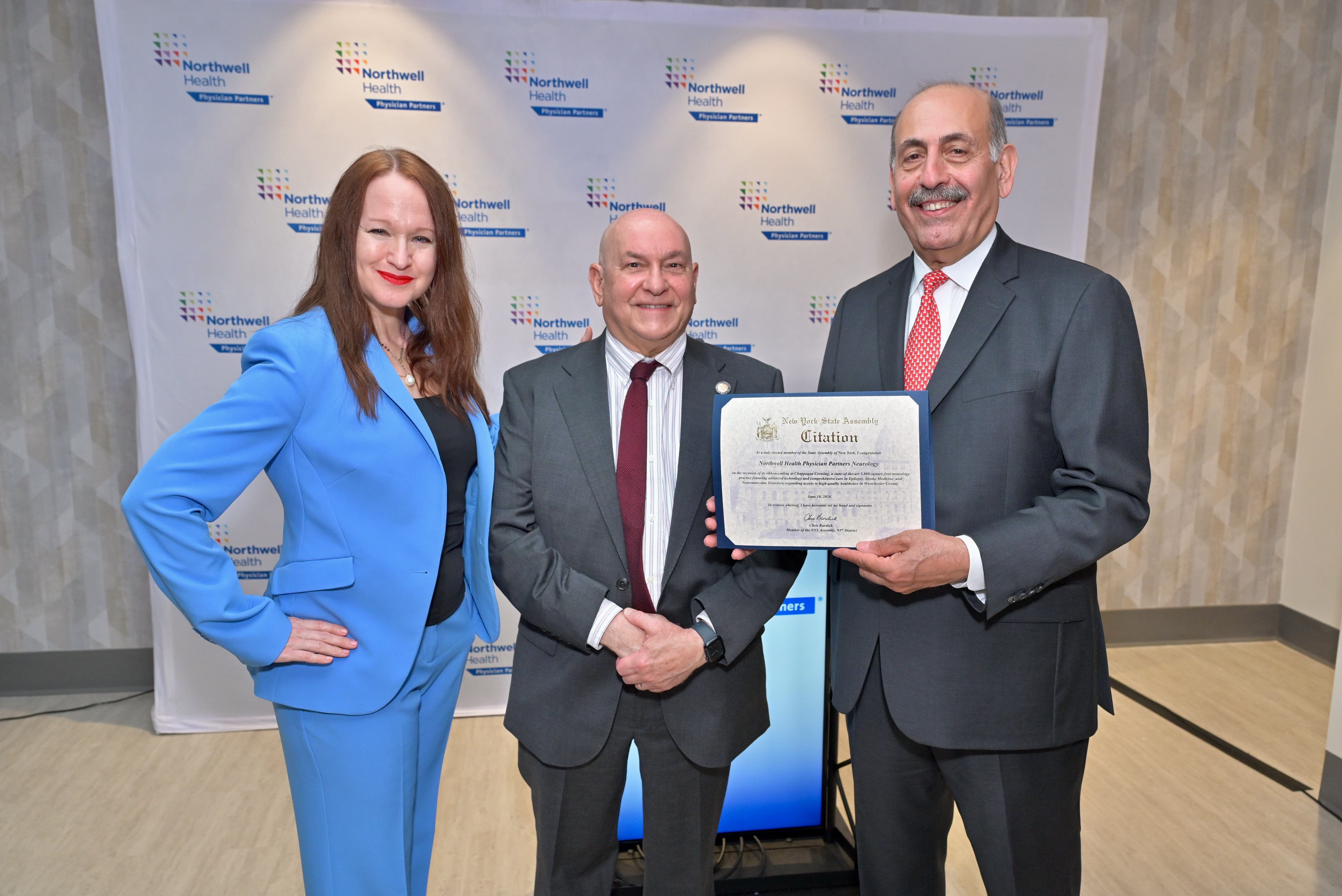 A woman in a powder blue suit holding her hands on her hips and two men in dark gray suits stand side by side with one holding up a citation.