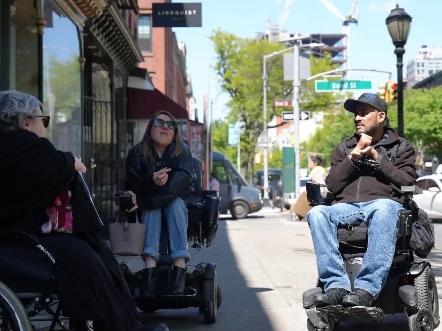 Nina Bakoyiannis and Jose Hernandez speak outside a Brooklyn district office during a disability rights rally on May 4, opposing limits on 24-hour home care shifts, as advocates call for changes to Intro. 303.