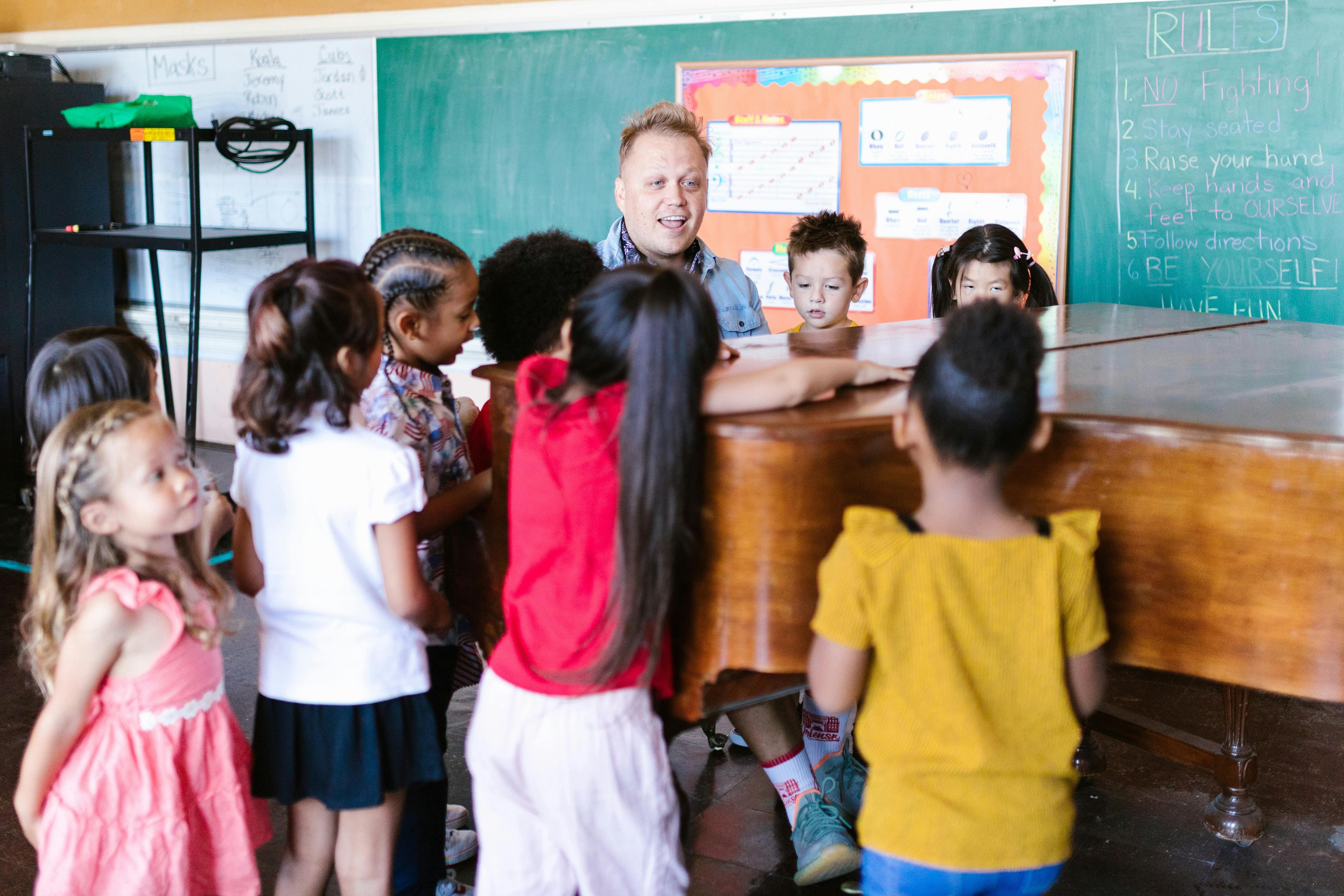 Students of different backgrounds in a classroom gathered around a piano while an instructor is playing.