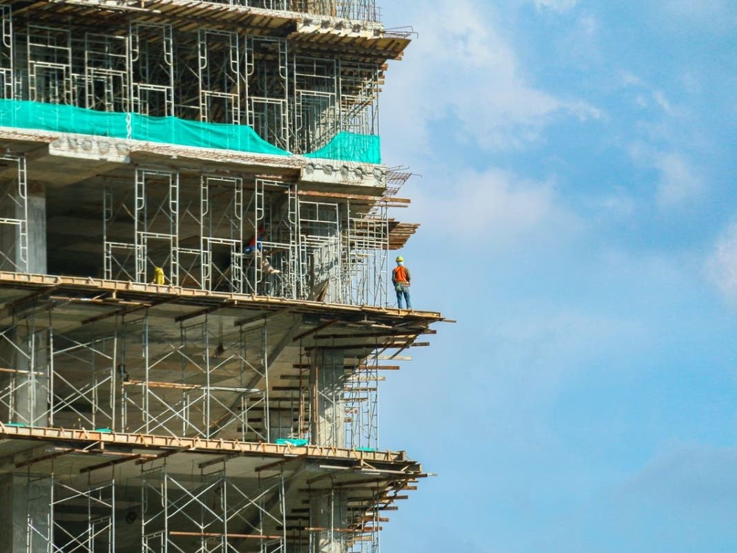 A construction worker stands on a scaffolding at a high-rise building under a blue sky.
