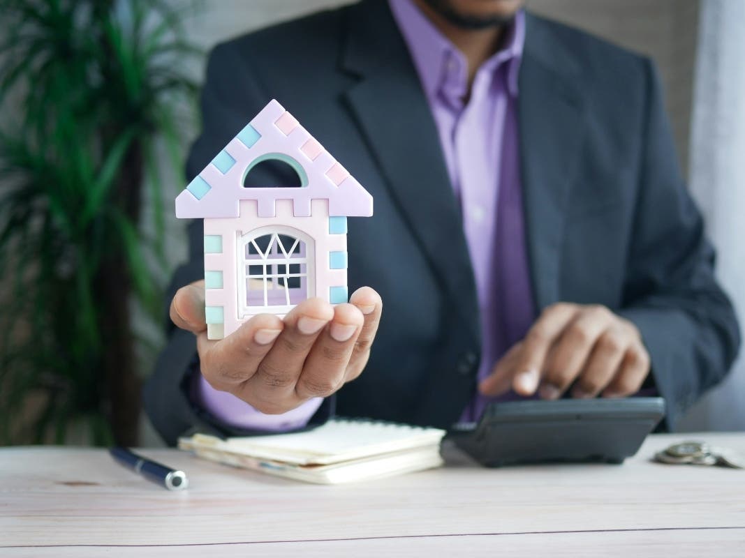 A man holding a model house and working on a calculator at his desk.
