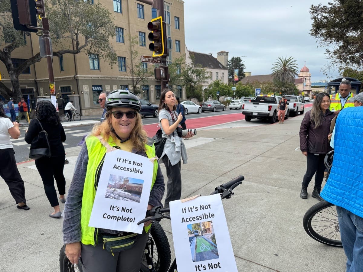 Helen Walsh, an adaptive cyclist, stands with her black three‑wheel cycle during a street-corner gathering, both sporting signs reading, “If It’s Not Accessible It’s Not Complete!”
