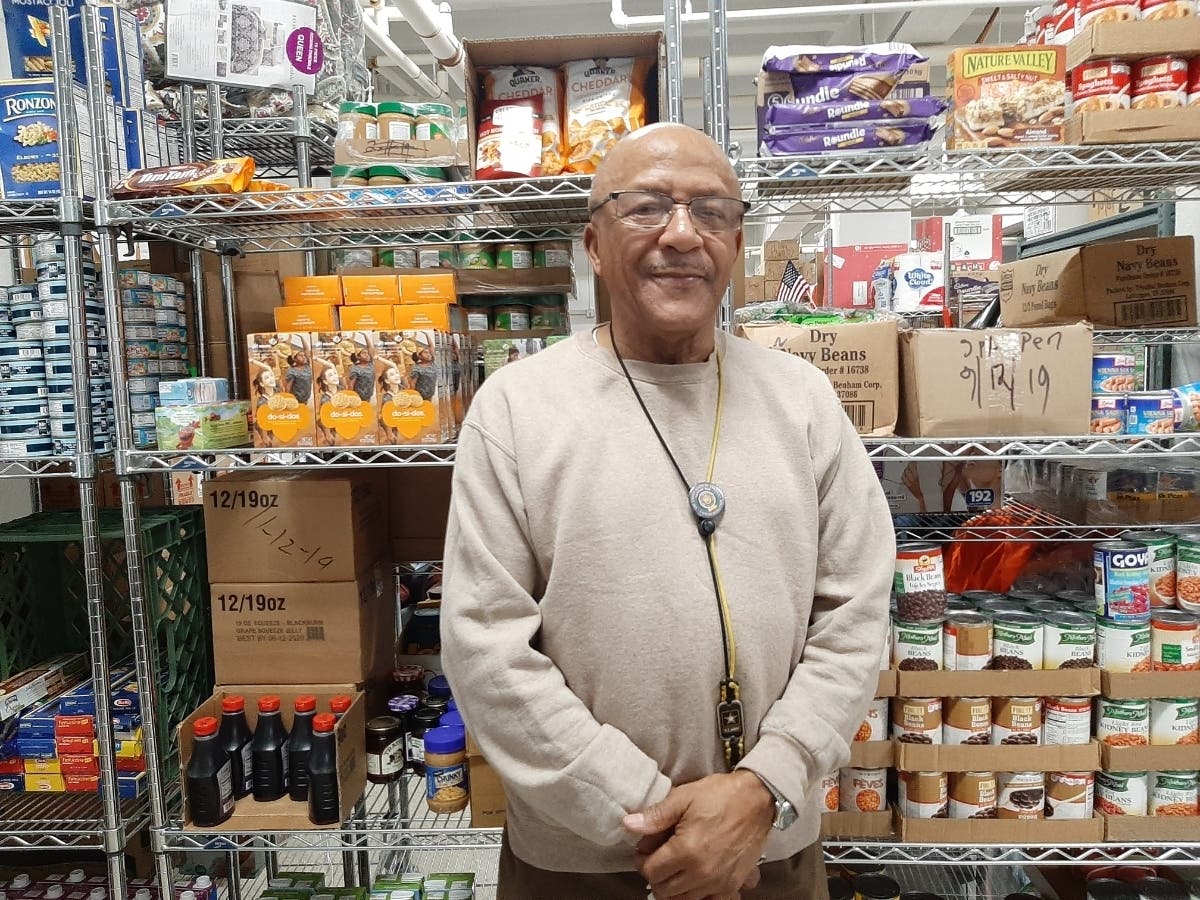 Montrose VA Food Pantry volunteer director, Duke Searles, poses in front shelf of goods that will benefit local Veterans in need. 